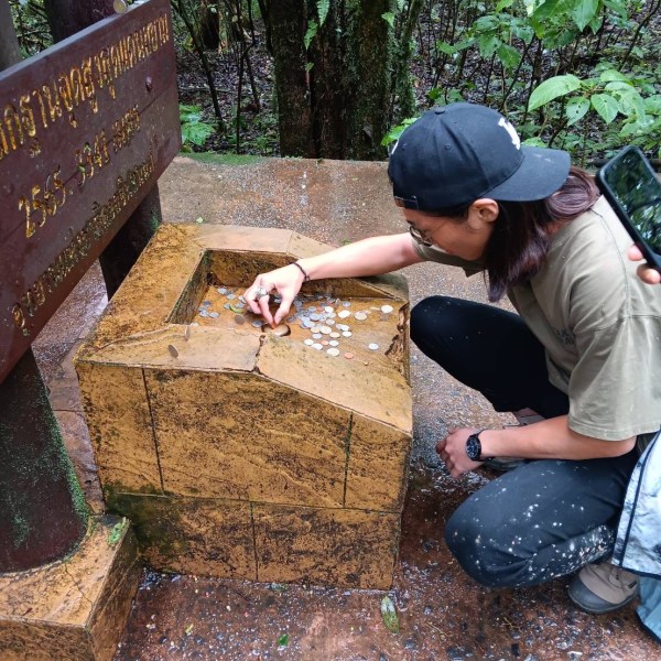 Person kneeling beside a stone monument with coins on top, in a forested area.