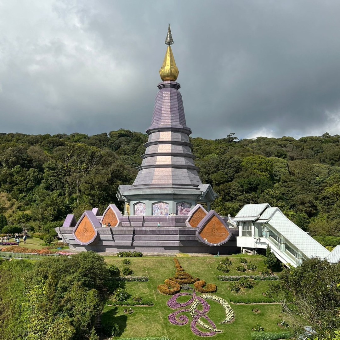 Ornate temple with golden spire, surrounded by garden, set against a cloudy sky and forested background.