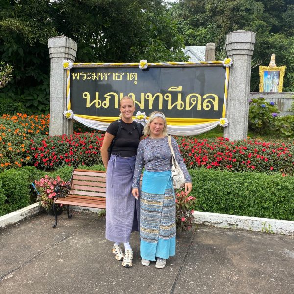 Two people posing in colorful attire in front of a sign with flowers and greenery.